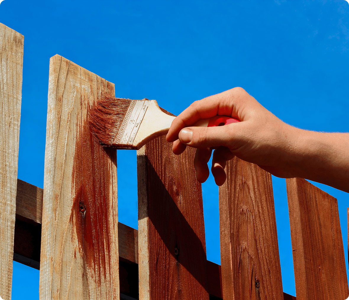 Hand painting wooden fence against blue sky.